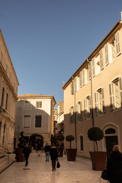 Sunny courtyard at Le Prieuré Notre-Dame in Montfrin with ochre buildings, shuttered windows, and pedestrians walking on