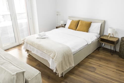Dormitorio luminoso en Ab Be Apartments Málaga con cama de matrimonio, almohadas doradas, mesita de noche de madera y vistas a la ciudad desde el balcón