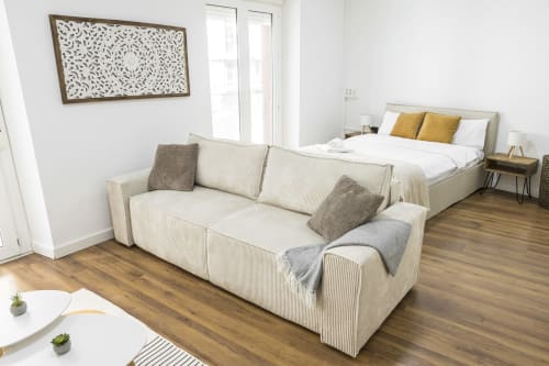 Estudio moderno en Ab Be Apartments, Málaga, con sofá beige, cama con cojines dorados, suelos de madera y obras de arte.