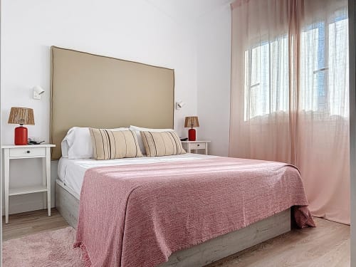 Dormitorio luminoso en la casa rural La Bella Lola en L'Estartit con cabecero beige, ropa de cama rosa y grandes ventanas con cortinas