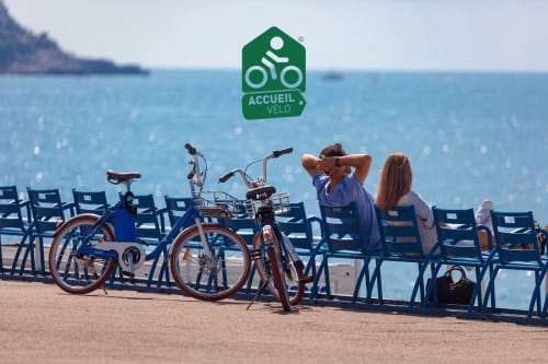 Ciclistas se relajan en sillas azules en la playa de Niza con el logo Accueil Vélo, Hôtel De La Fontaine