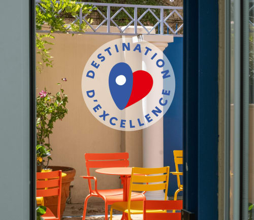 La entrada del patio del Hôtel De La Fontaine Nice cuenta con un letrero 'Destination D'Excellence' y sillas de colores.