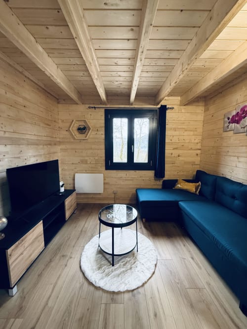 Salon chaleureux avec plafond à poutres apparentes, canapé bleu, téléviseur et table ronde aux Cabanes du Belvédère