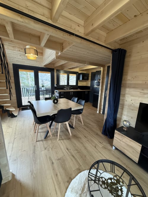 Intérieur de lodge moderne avec poutres en bois, table à manger, chaises noires, cuisine ouverte et terrasse avec vue sur les montagnes