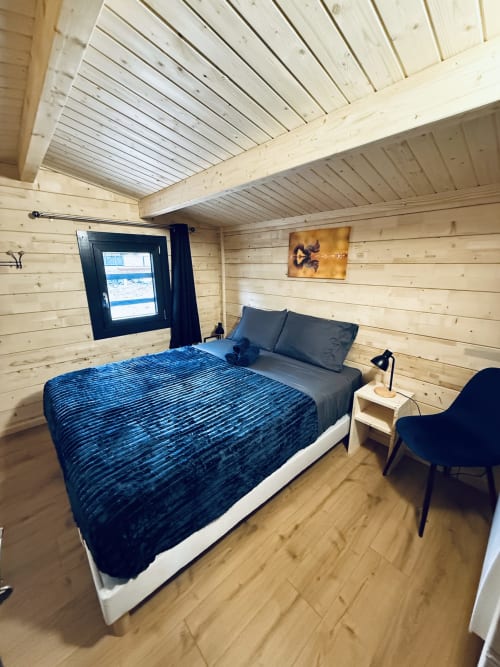 Chambre de lodge rustique avec lit queen-size, linge de lit bleu, plafond à poutres apparentes, baie vitrée vue sur le lac