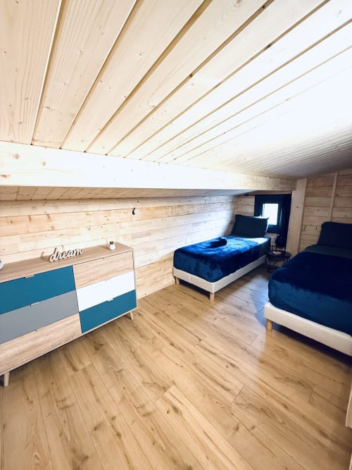 Chambre de lodge moderne aux poutres en bois, deux lits bleus et lumière naturelle aux Cabanes du Belvédère