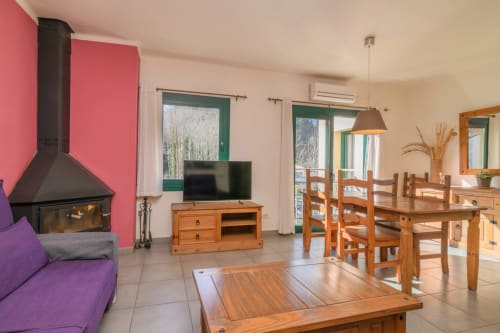 Amplia sala de estar con chimenea, sofá púrpura, mesa de comedor de madera, televisión y ventanas con marco verde con vistas