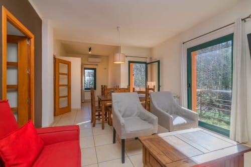 Sala de estar de cabaña moderna con sofá rojo, sillones grises, mesa de comedor de madera y puertas verdes hacia una terraza con vistas a las montañas