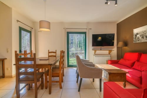 Open-plan living and dining area in Rural Montseny cottage with red sofa, wooden table, and mountain views through green