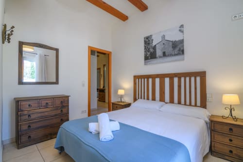 Bright bedroom in Rural Montseny cottage with wooden bed, white linens, blue runner, nightstands with lamps, dresser with