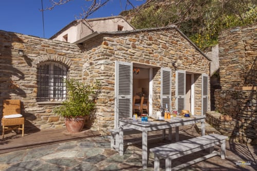 Cour de villa en pierre avec table à manger, portes volets, plantes en pots et fenêtre cintrée à La Conciergerie du Cap Corse