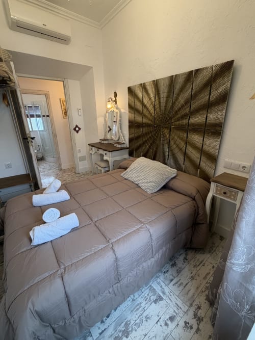 Dormitorio con cama acolchada marrón, toallas blancas, cabecero de panel decorativo de madera y baño en suite en casita de Almagro