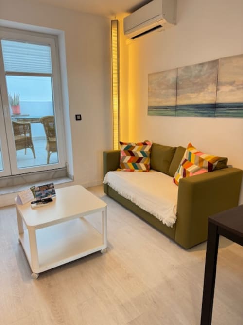 Acogedora sala de estar con sofá verde oliva, mesa de café blanca, cojines coloridos y balcón con vista al mar en El Faro De