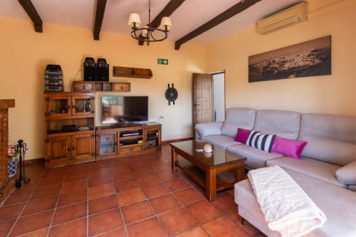 Spacious living room at Finca Navalpino with terra cotta floors, wooden beams, gray sofa, and wooden entertainment center in