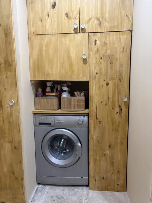 Buanderie intégrée avec meubles en bois, machine à laver grise et paniers de rangement dans l'appartement d'Arles.