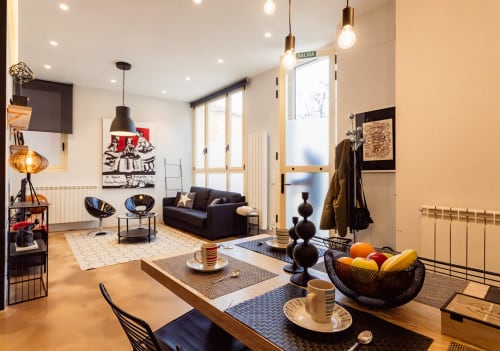 Moderno loft en Burgos con sala abierta, comedor, sofá negro, lámparas colgantes y vistas a la ciudad desde el balcón