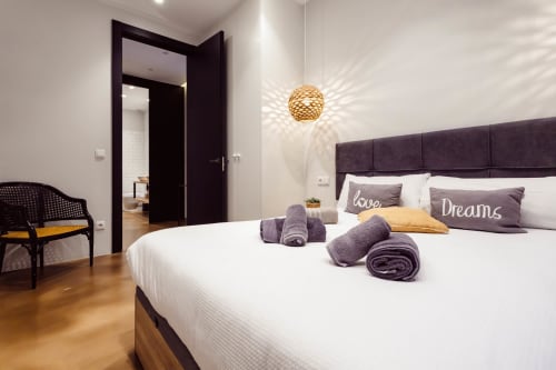 Moderno dormitorio en Loft Center Burgos con cama blanca, cabecero morado, lámpara colgante dorada y puerta de entrada oscura