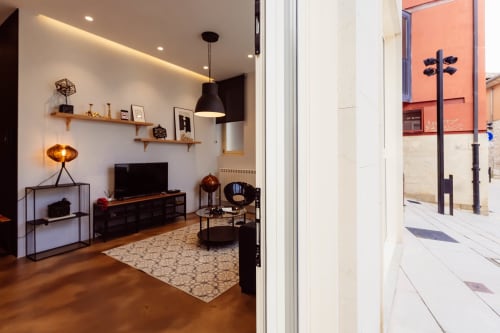 Sala de estar de loft moderno en Burgos con suelos de madera, TV, estanterías, iluminación colgante y vistas a la ciudad a través de cristal