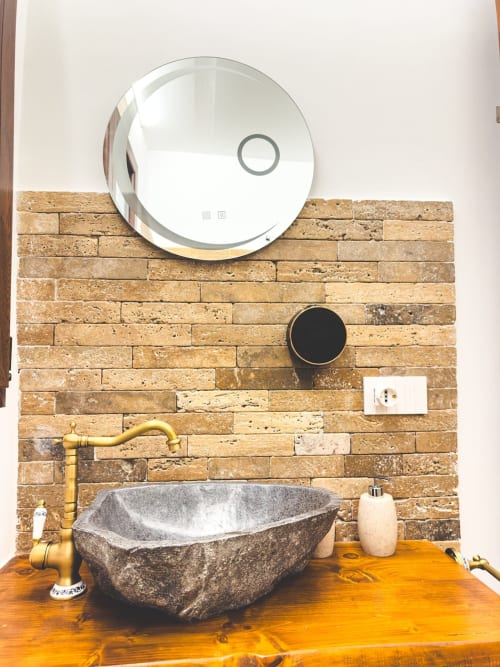 Lavabo moderno con ciotola in pietra, rubinetteria in ottone, specchio rotondo e parete in laterizio presso CorteLirice Paradiso