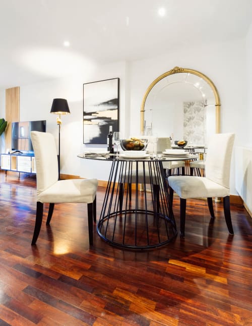 Comedor moderno en apartamento de Burgos con mesa redonda negra, sillas crema, espejo dorado y suelos de madera