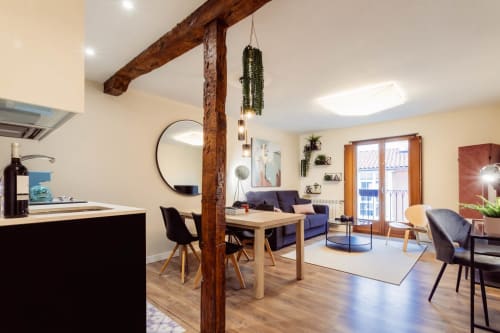 Moderno salón en Burgos con vigas de madera, mesa de comedor, sofá azul marino y vistas a la ciudad desde el balcón