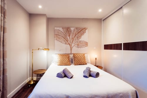 Dormitorio moderno en The Charm of Twelve de Burgos con cama blanca, almohadas doradas, toallas enrolladas, cuadro de plantas y madera