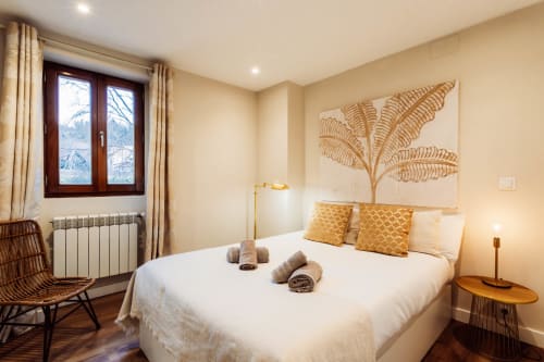 Moderno dormitorio en Unique Rooms Burgos con cama blanca, cojines dorados, arte botánico en la pared y vistas a la ciudad desde el balcón