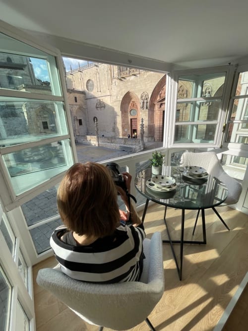 Mujer fotografiando la Catedral de Burgos desde apartamento moderno con ventanales de vidrio, mesa pulida y asientos con rayas.