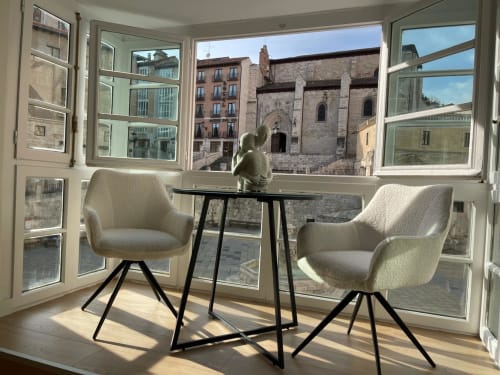 Apartamento moderno en Burgos con dos sillas color crema, mesa negra y vistas a la catedral a través de amplios ventanales