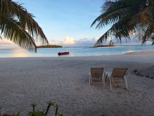 Tropical beach at Tikehau with two loungers, red boat, palm fronds, and calm turquoise waters at sunset