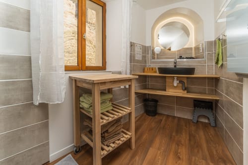 Salle de bain moderne du gîte Au Château De Villelongue avec meuble-lavabo en bois, miroir rond et accents en pierre