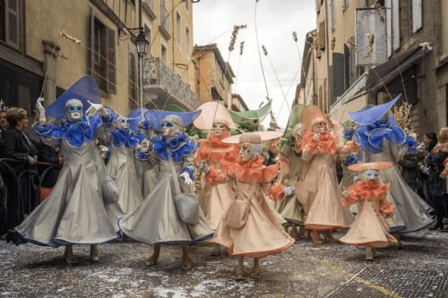 Procesión del carnaval veneciano en Villelongue-d'Aude con artistas disfrazados luciendo máscaras azules y naranjas con elaboradas alas