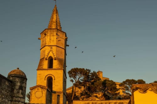 Golden-hour view of historic church bell tower at Le Prieuré Notre-Dame in Montfrin, France with fortress walls and birds