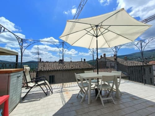 Terrazza soleggiata con ombrellone bianco e tavoli da pranzo con vista sul castello di Monteriggioni e la campagna toscana