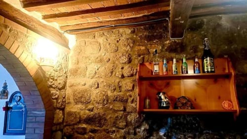 Interno rustico in pietra con travi in legno, scaffali arancioni che espongono bottiglie e oggetti decorativi a Torre Della