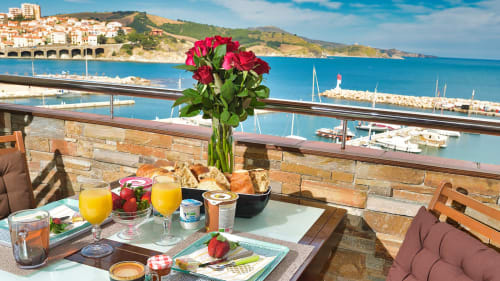 Table de petit-déjeuner avec vue sur le port de Banyuls-sur-Mer, roses rouges, viennoiseries fraîches, jus et mer Méditerranée