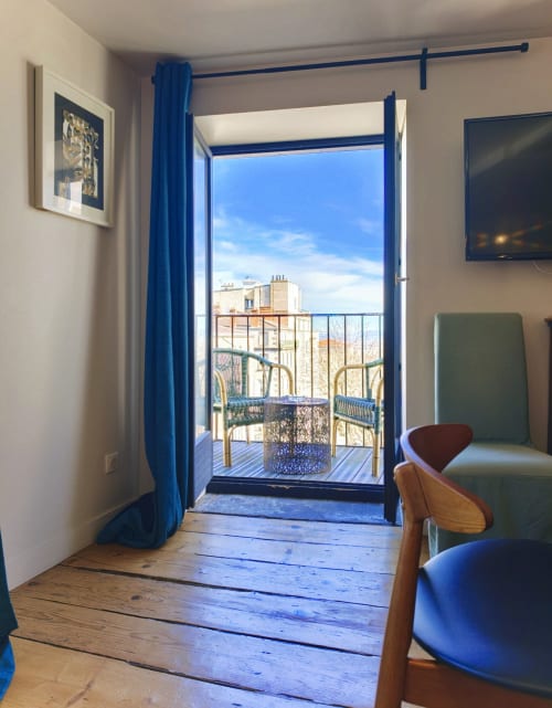 Chambre d'hôtel lumineuse à Clermont-Ferrand avec rideaux bleus, parquet et balcon ouvert sur la skyline