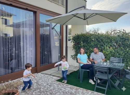 A family with children relaxing on the outdoor patio of Casa Meravigliosa in Fontanafredda, furnished with a garden table, a gray umbrella, and a manicured hedge.