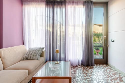 "Bright living room at Casa Meravigliosa in Fontanafredda with beige sofa, glass coffee table, mosaic floor, and large curtains on the French window overlooking the garden."