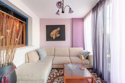 "Relaxation area at Casa Meravigliosa in Fontanafredda with a large beige corner sofa, a glass coffee table, lilac walls with decorative paintings, and a matching purple chandelier."