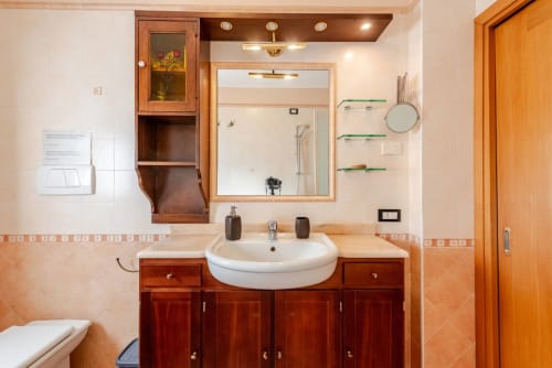 "Detail of the sink area at Casa Meravigliosa in Fontanafredda with dark wood vanity, light marble countertop, large illuminated mirror, and glass shelves."