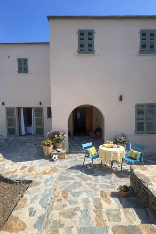 Cour ensoleillée de la Villa Marine à Centuri avec chaises bleues, jardinières fleuries et terrasse en pierre menant à l'entrée voûtée