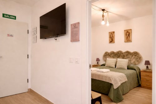 Entrada del apartamento en Mérida con TV y vista al dormitorio con ropa de cama verde y mesas de noche de madera