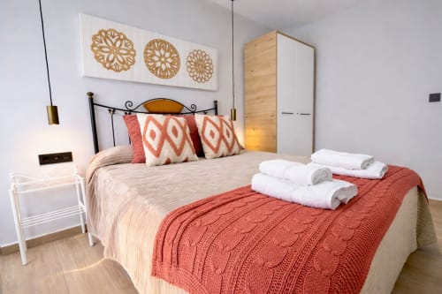 Dormitorio moderno en apartamento de Mérida con almohadas estampadas rojas, toallas enrolladas, armario de madera y arte geométrico en pared.