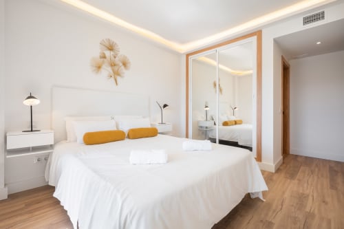 Modern bedroom in Marbella apartment with white bed, golden accents, mirrored closet, and wooden flooring