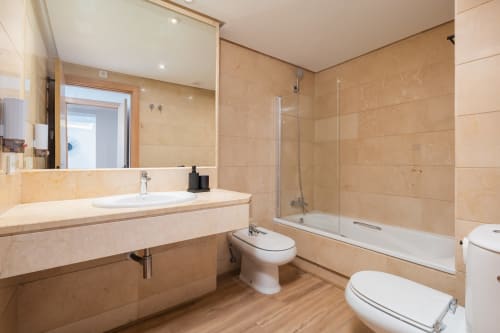 Modern bathroom in Marbella apartment with beige tile, vanity sink, glass shower enclosure, and two toilets