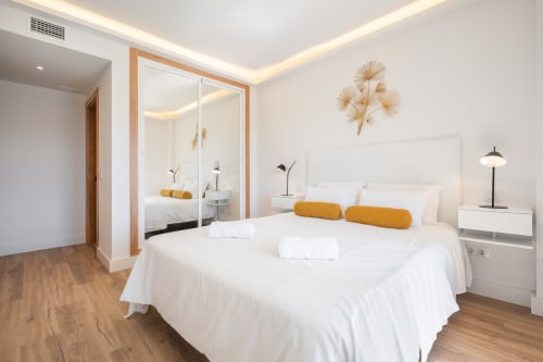 Modern bedroom in Marbella apartment with white bed, gold accents, mirrored wardrobe, wooden floors, and city view