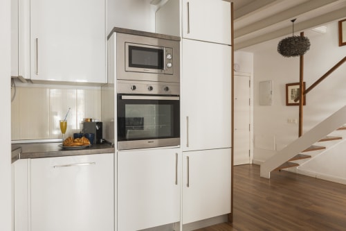 Cocina moderna blanca con electrodomésticos de acero inoxidable, encimera gris y escaleras de madera en el ático Ab Be, Málaga