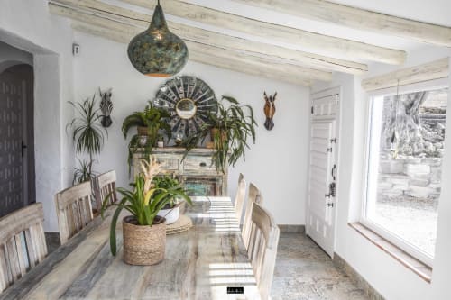 Rustic Spanish hotel dining room with wooden table, potted plants, decorative pendant lamp, and terracotta-tiled floor