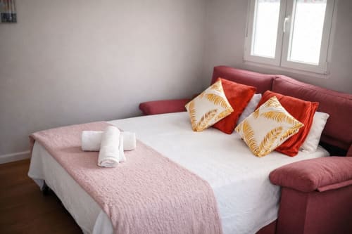 Dormitorio luminoso en Mérida con cama blanca, cabecera borgoña, almohadas decorativas, manta rosa y toallas enrolladas